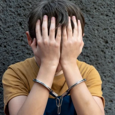 shutterstock_2279885581 A 10-year-old boy in handcuffs sits on a gray background.