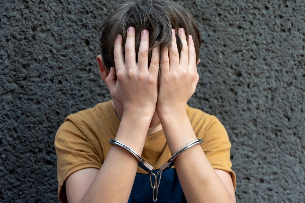 A 10-year-old boy in handcuffs sits on a gray background.